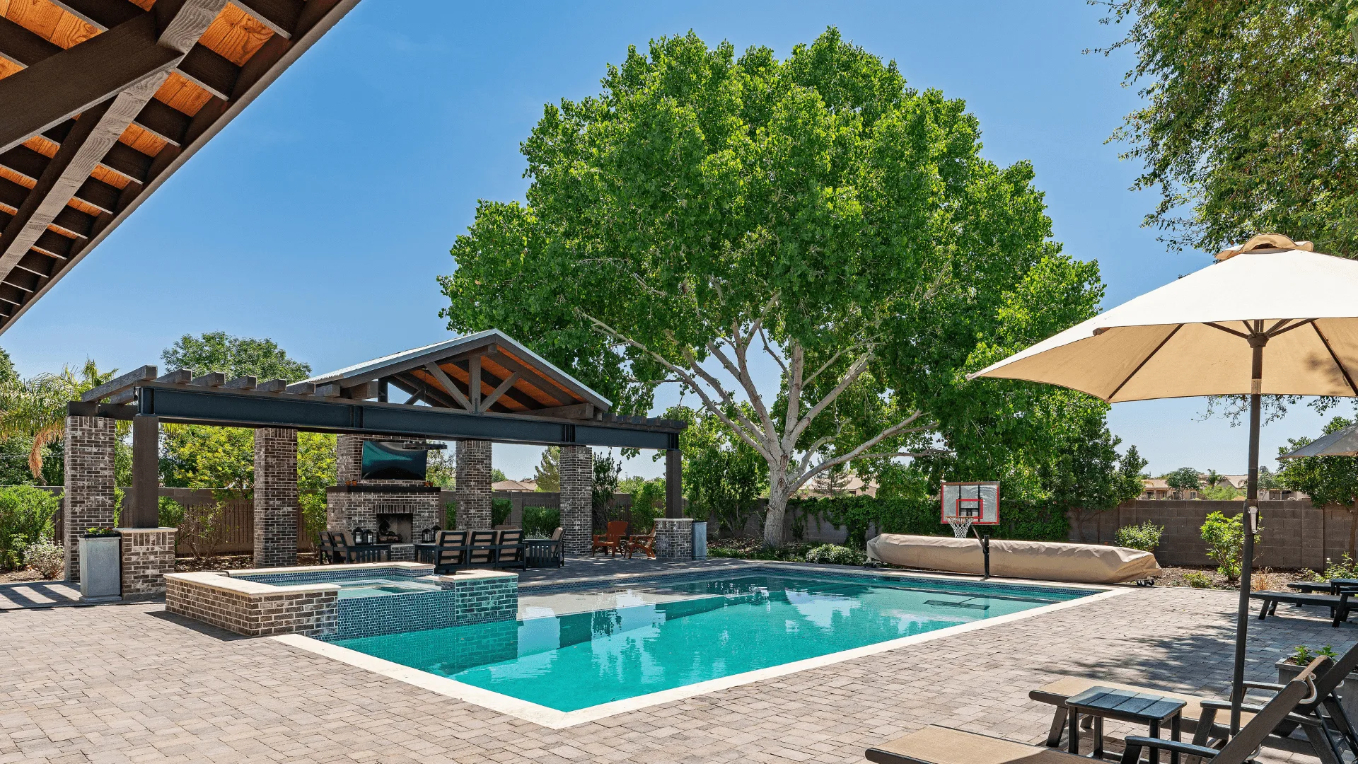 fiberglass swimming pool with a stone deck surrounded by seating and a shaded area featuring a large tree in the background